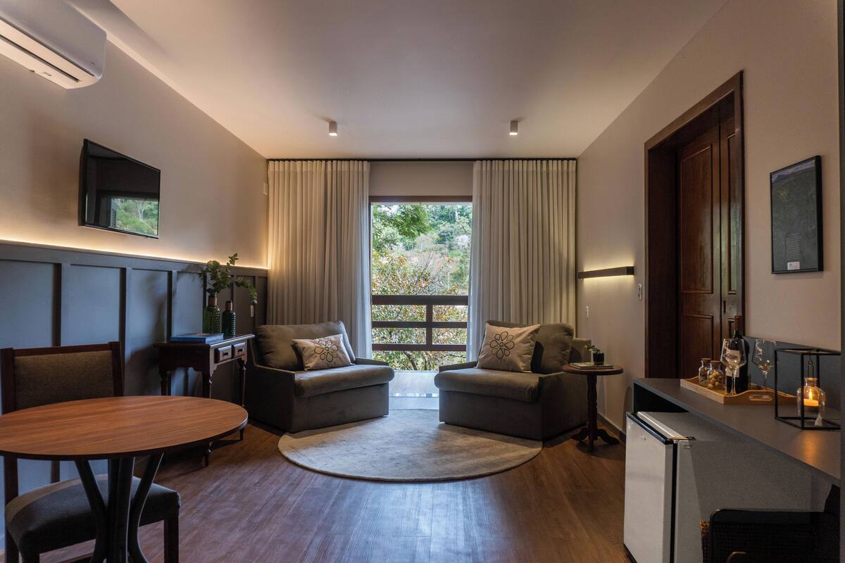 A modern living room with a dining table, two sofas, a small kitchen area, and a large window with curtains.