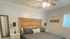 Een slaapkamer met een houten hoofdbord, een bed met grijze beddengoed, een nachtkastje met een lamp, een plafondventilator, een spiegel en een bord aan de muur.