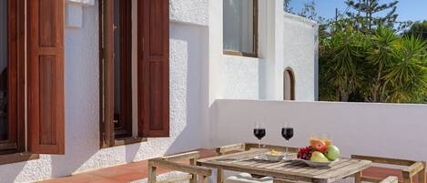 A wooden table with two wine glasses and a bowl of fruit on a terrace with white walls and wooden shutters.