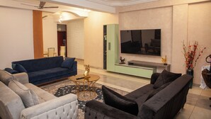 A modern living room with a blue sofa, a black sofa, and a beige armchair. There is a TV mounted on the wall, a ceiling fan, and a decorative vase with branches. - Mercy Royal Heights (Kumasi)
