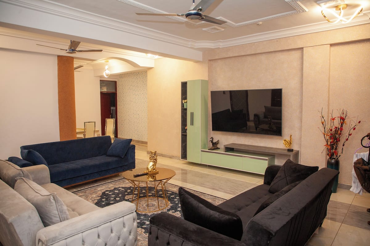 A modern living room with a blue sofa, a black sofa, and a beige armchair. There is a TV mounted on the wall, a ceiling fan, and a decorative vase with branches.