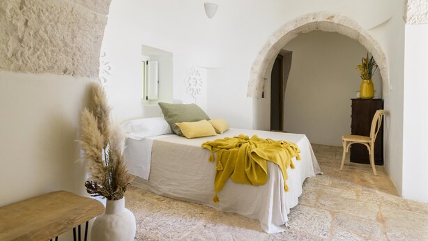 A bedroom with a bed, a chair, a wooden bench, a vase with dried plants, and an arched doorway.