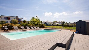 En swimmingpool med træterrasse, omgivet af liggestole og et indhegnet område med huse i baggrunden.