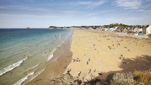 En strand med folk, der solbader og svømmer, en klippefyldt kystlinje og bygninger i baggrunden.