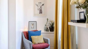 A living room with a grey armchair, a red cushion, a framed picture on the wall, a potted plant, and a shelf with a vase of flowers. - Live In The Heart Of Bustling Camden (London)