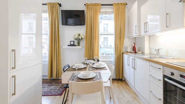 A modern kitchen with white cabinets, a wooden countertop, and a dining area with a table set for four. - Live In The Heart Of Bustling Camden (London)