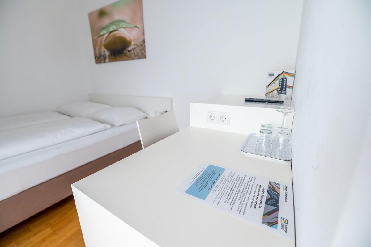 A clean, modern hotel room with a single bed, a small desk with a computer and glasses, and a wall-mounted power outlet.