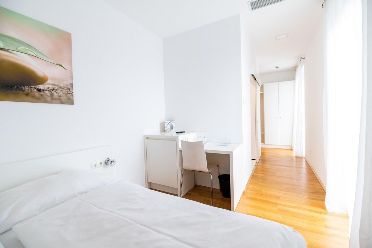 A bedroom with a bed, a desk, and a chair. There is a painting on the wall and a closet in the background.