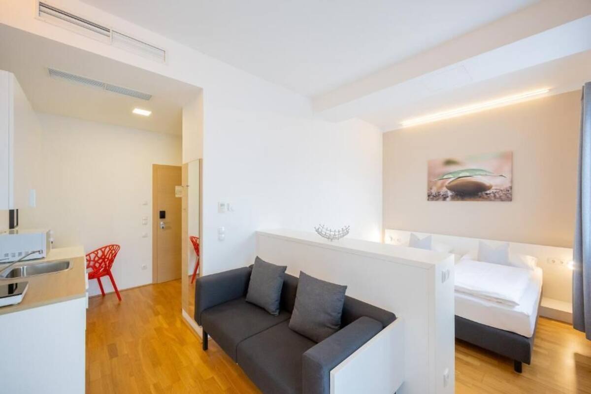 A modern hotel room with a sofa, a bed, a small kitchen area, and a red chair.
