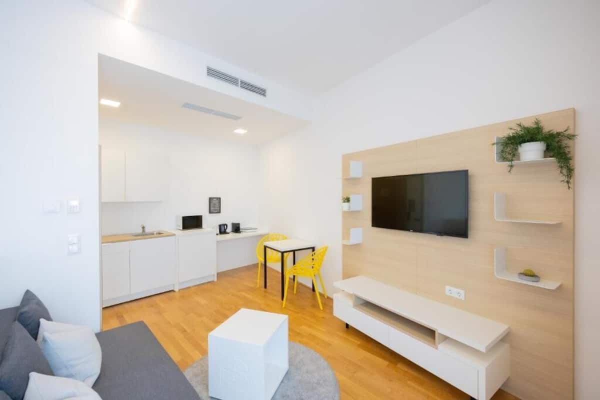 A modern living room with a flat-screen TV, a sofa, and a dining area with yellow chairs.