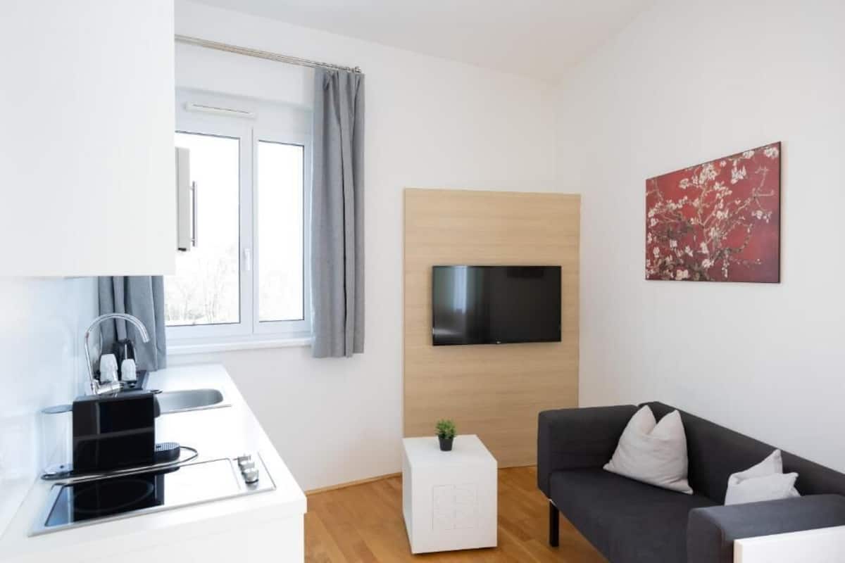 A modern living room with a flat-screen TV, a dark sofa, a small white side table, and a window with curtains.