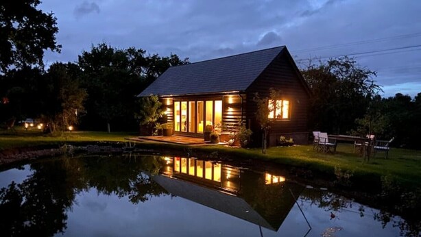 A lodge with a pond in front, wooden exterior, and a well-lit porch. - Stunning Lodge in Idyllic Setting, Near Cambridge (Bourn)
