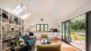 A spacious living room with a sofa, coffee table, and a dining area with chairs. There is a kitchen in the background with a window overlooking a garden. - Stunning Lodge in Idyllic Setting, Near Cambridge (Bourn)