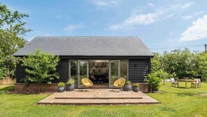 A modern house with a dark exterior, a covered patio with two chairs, and a lawn. - Stunning Lodge in Idyllic Setting, Near Cambridge (Bourn)