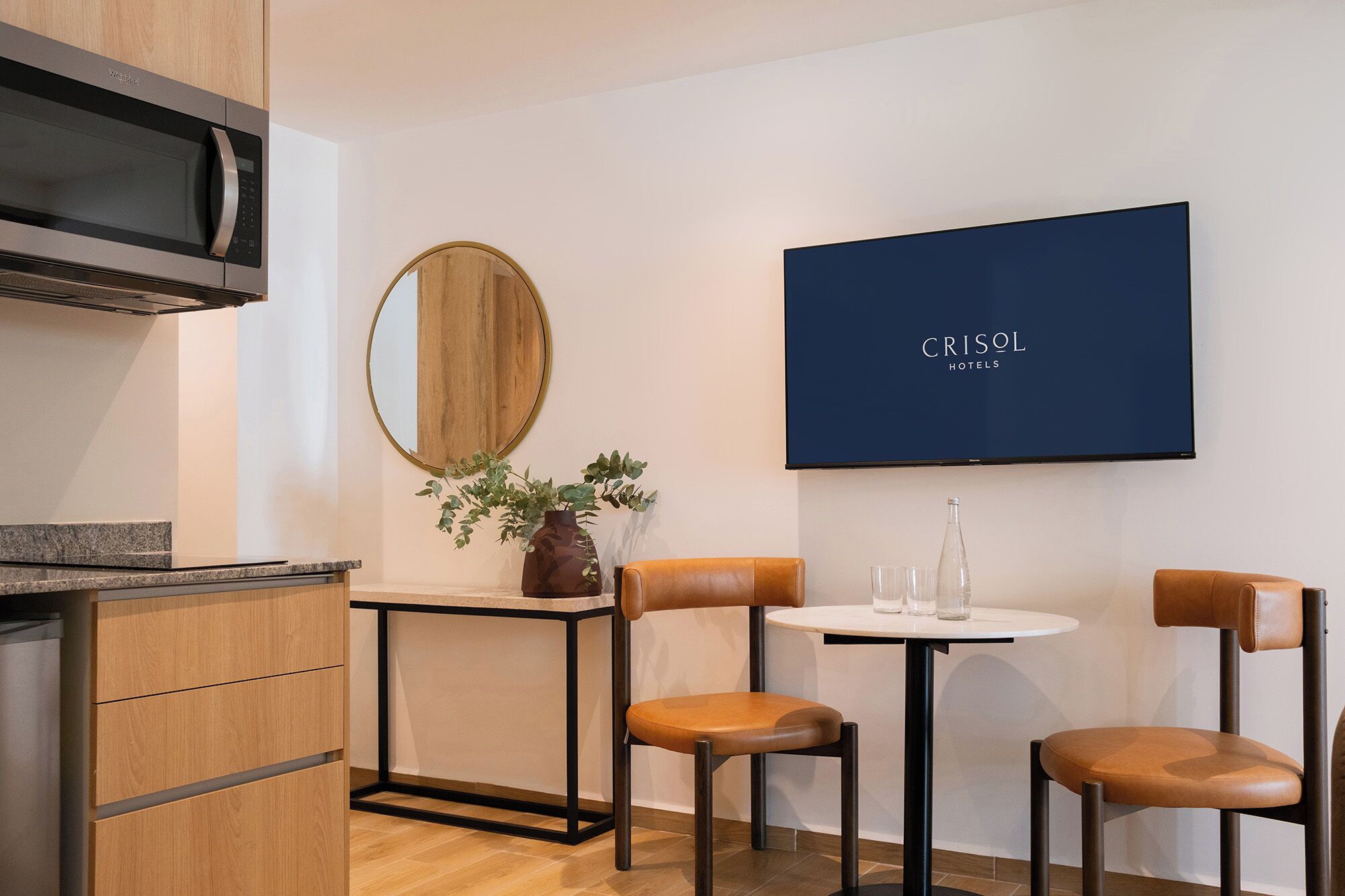 Een moderne keuken met een magnetron, houten kastjes, een ronde spiegel, een potplant en een tafel met twee stoelen.