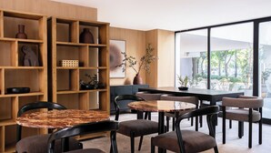A modern interior with wooden shelving, a dining area with round tables and chairs, and a large window offering a view of greenery. - Crisol Suites Condesa (Mexico City)