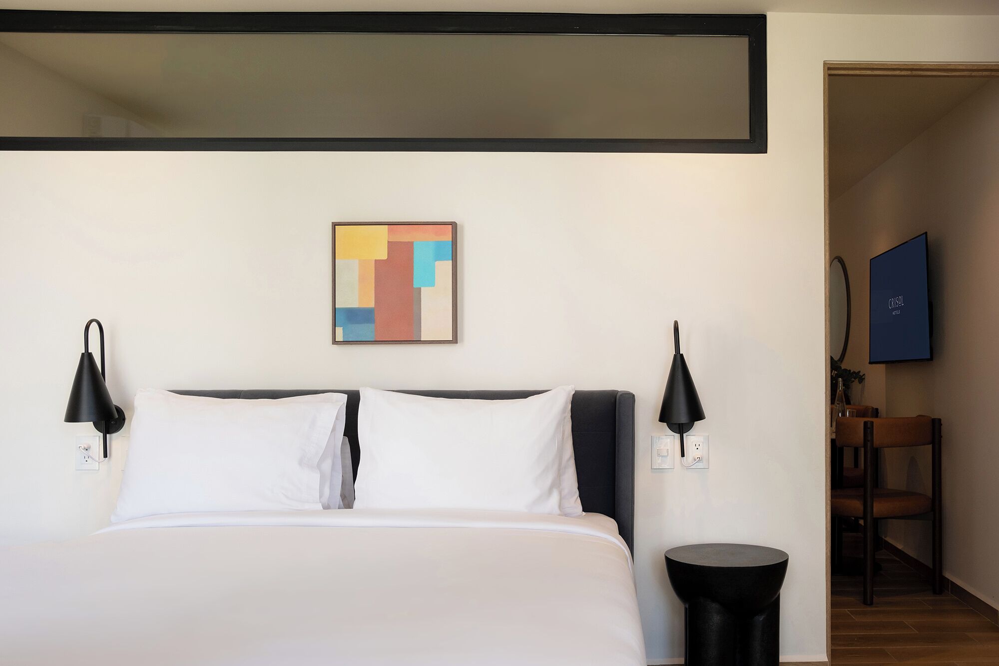 A hotel room with a bed, two bedside lamps, a small round table, a chair, and a painting on the wall.