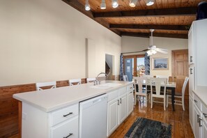 A modern kitchen with white cabinetry, a central island with a sink, and a dining area with a table and chairs. - Moonshadow: Oceanfront, walkway to the beach, hot tub, awesome ocean views! (Nags Head)