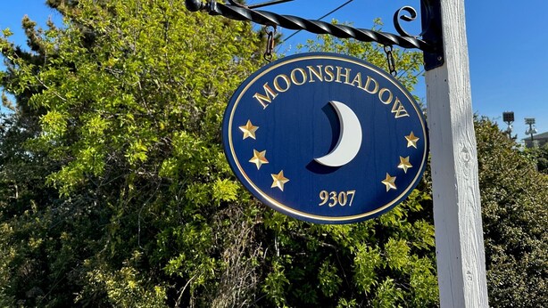 A sign with the text "MOONSHADOW" and the number "9307" against a backdrop of trees and a clear sky. - Moonshadow: Oceanfront, walkway to the beach, hot tub, awesome ocean views! (Nags Head)