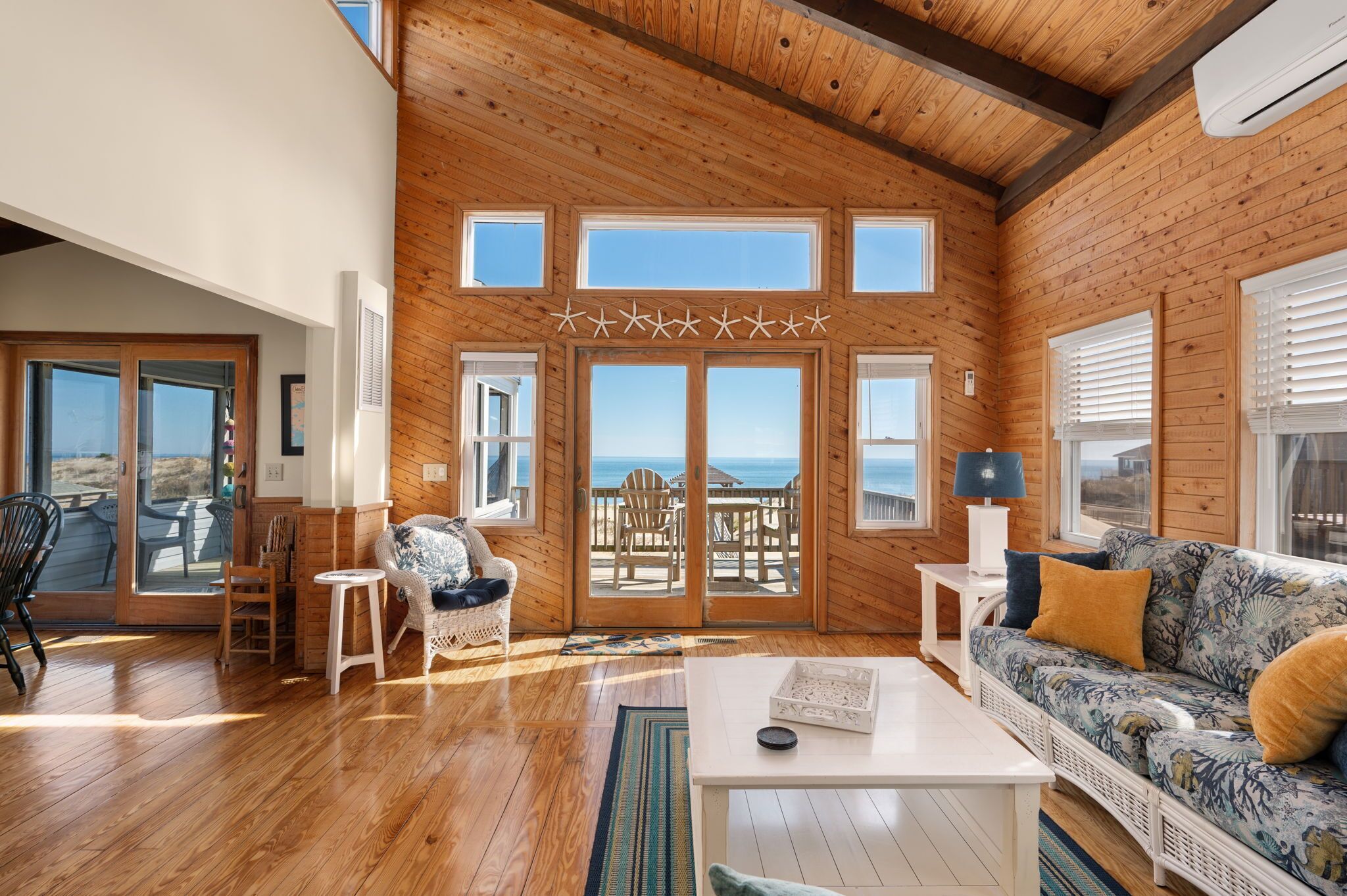 A spacious living room with wooden floors, a white sofa, a wicker chair, and a view of the beach through large windows.