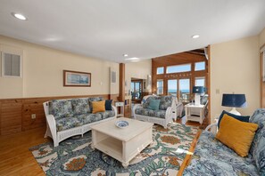 A cozy living room with wicker furniture, a patterned rug, and a view of the sea through large windows.
