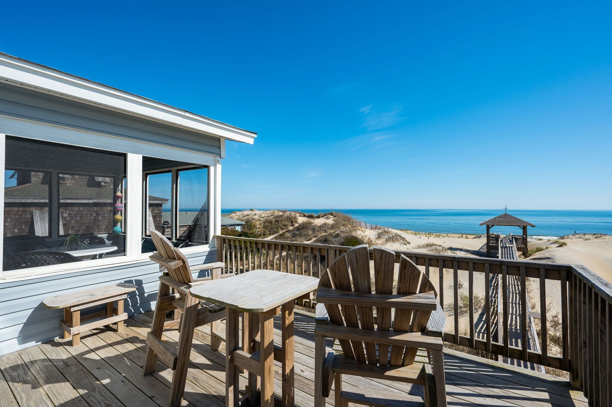 Una terraza de madera con una mesa y sillas, con vista a una playa con dunas y el océano.