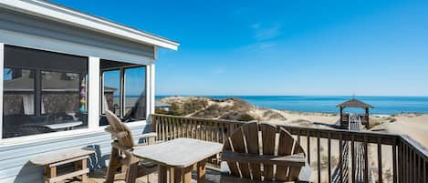 Una terraza de madera con una mesa y sillas, con vista a una playa con dunas y el océano.