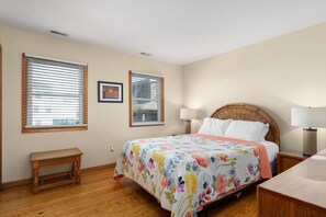 A bedroom with a bed, two bedside lamps, a bench, and two windows with blinds.