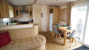 A compact kitchen with a dining area, a sofa, and a view through a large window. - 3 Bedroom Caravan Pets Free Linen Included (King's Lynn)