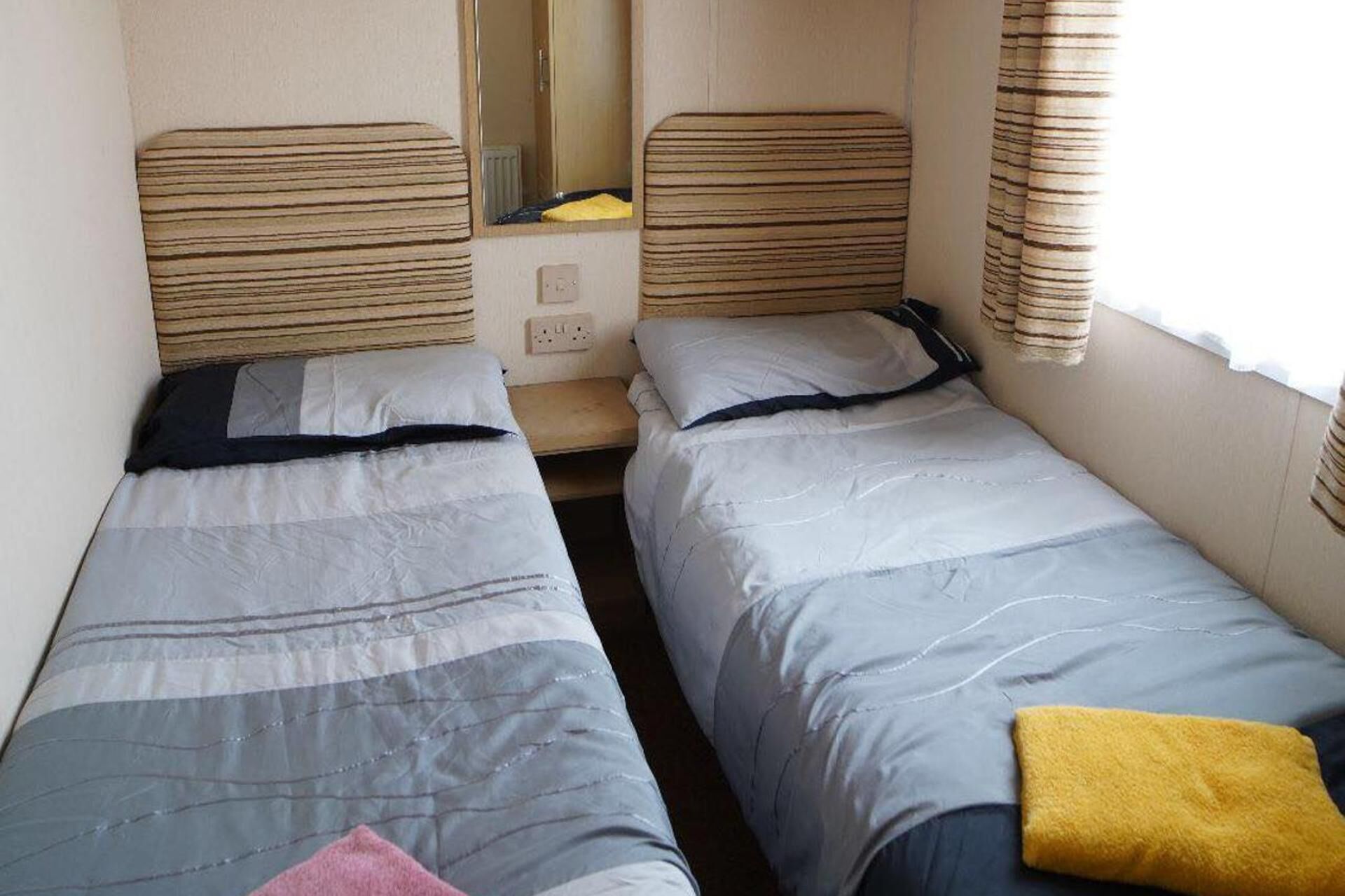 A small, compact room with two single beds, a mirror, and a window with curtains.