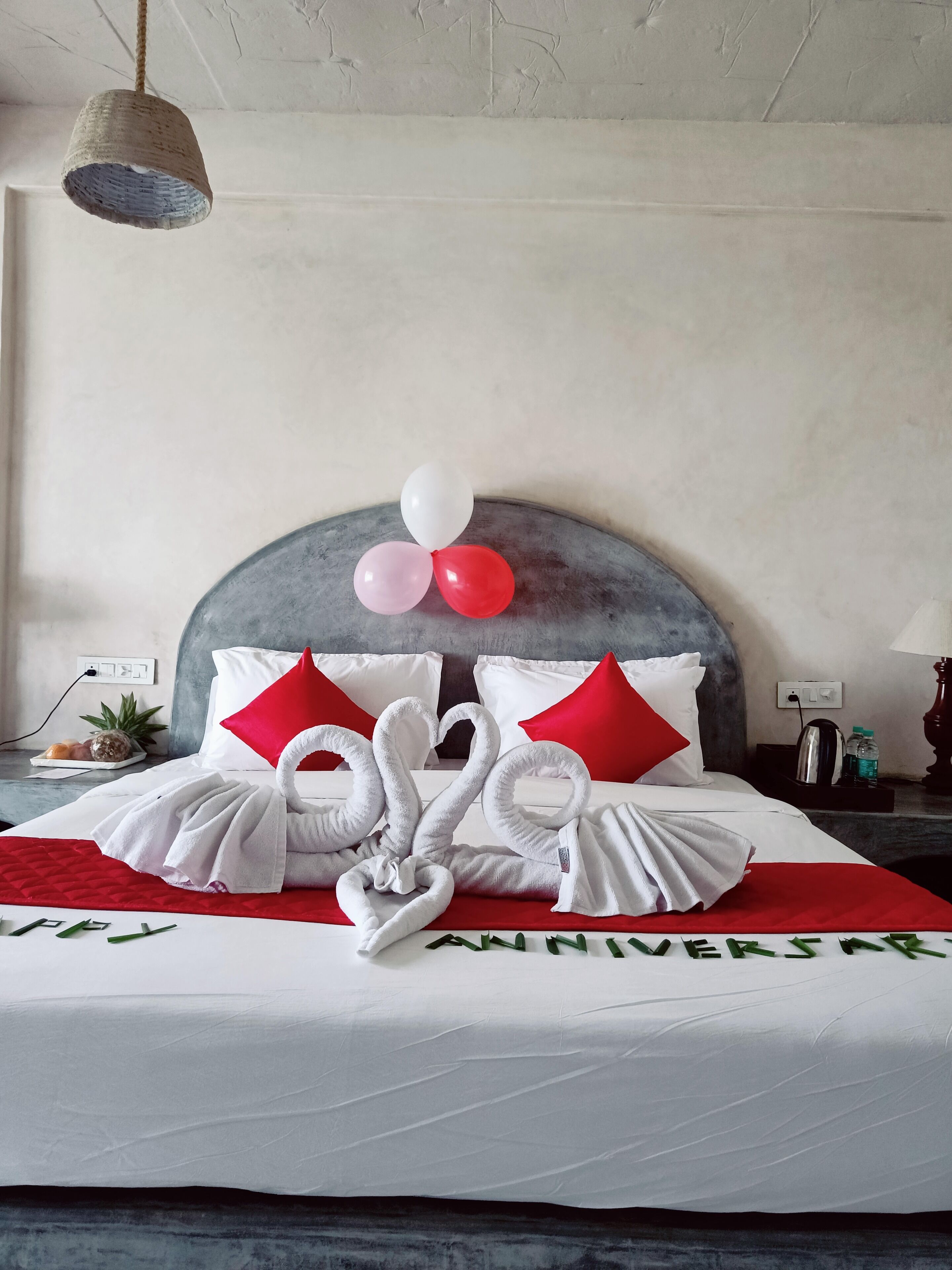 A neatly made bed with white and red pillows, a heart-shaped towel arrangement, and a 'Happy Anniversary' sign.
