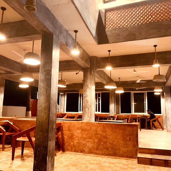 A spacious restaurant with high ceilings, exposed beams, and pendant lighting. Tables are arranged for dining, and there are large windows providing natural light.