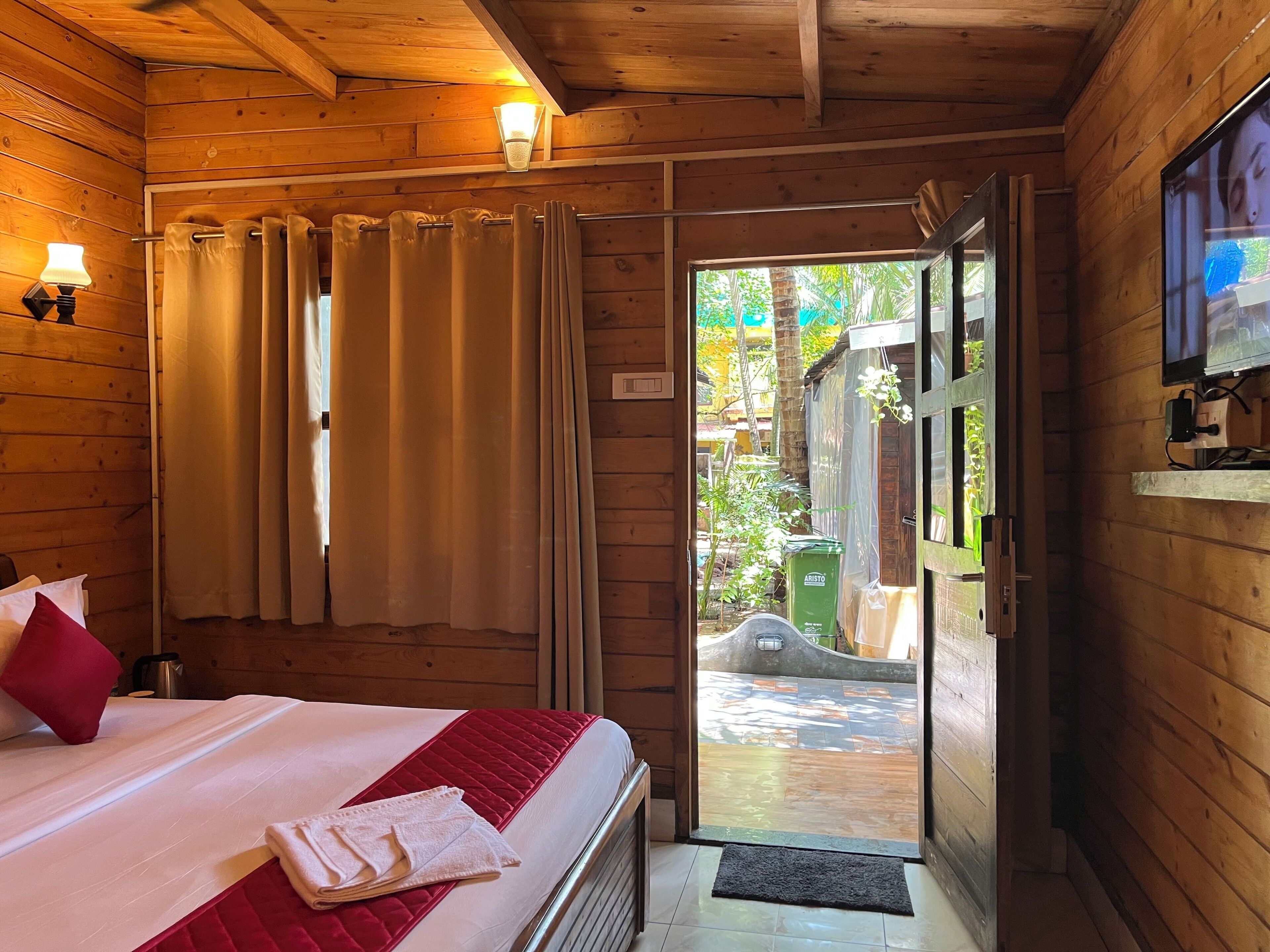 A wooden cabin room with a bed, curtains, a television, and a view of the outside.