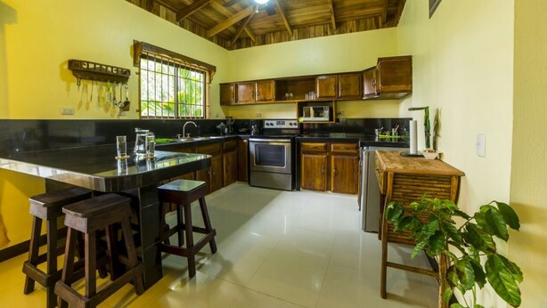 Una cocina con armarios de madera, una isla central con taburetes, un ventilador de techo y una ventana con rejas.