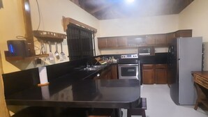 Una cocina compacta con encimera negra, armarios de madera, microondas, refrigerador y una mesa de madera.
