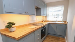 Une cuisine moderne avec des meubles gris, un plan de travail en bois, un four encastré et une fenêtre avec des stores.