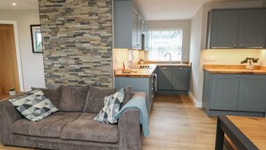 Un espace de vie compact comprenant un canapé gris, un mur en pierre, une petite cuisine avec des meubles gris et un plancher en bois.