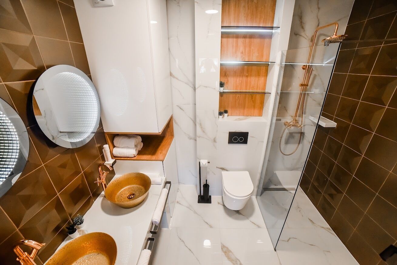 A modern bathroom with a marble floor, a wooden vanity, a round sink with a copper faucet, a toilet, and a glass shower enclosure.