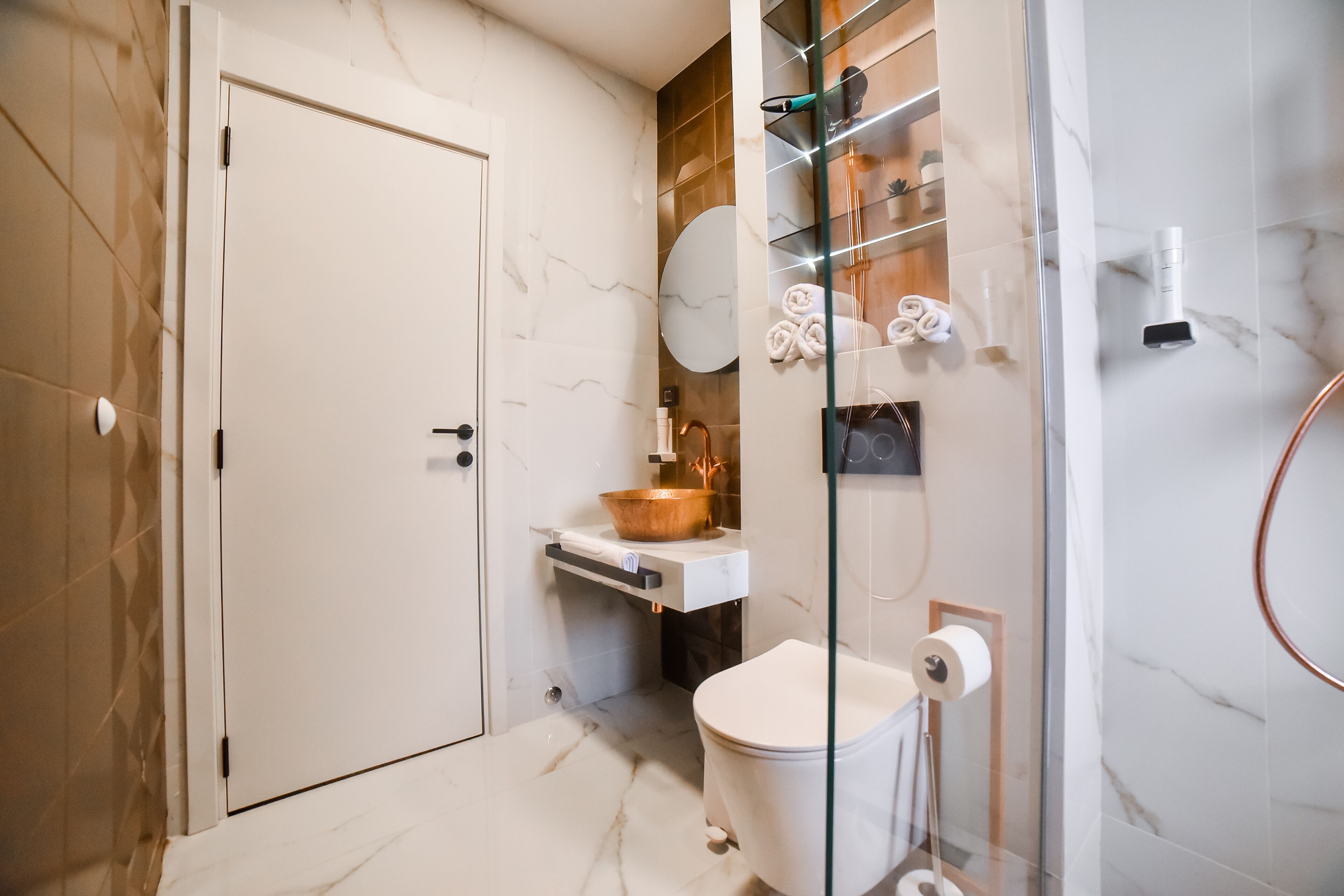 A bathroom with a white door, a copper sink, a toilet, and a glass shower enclosure.