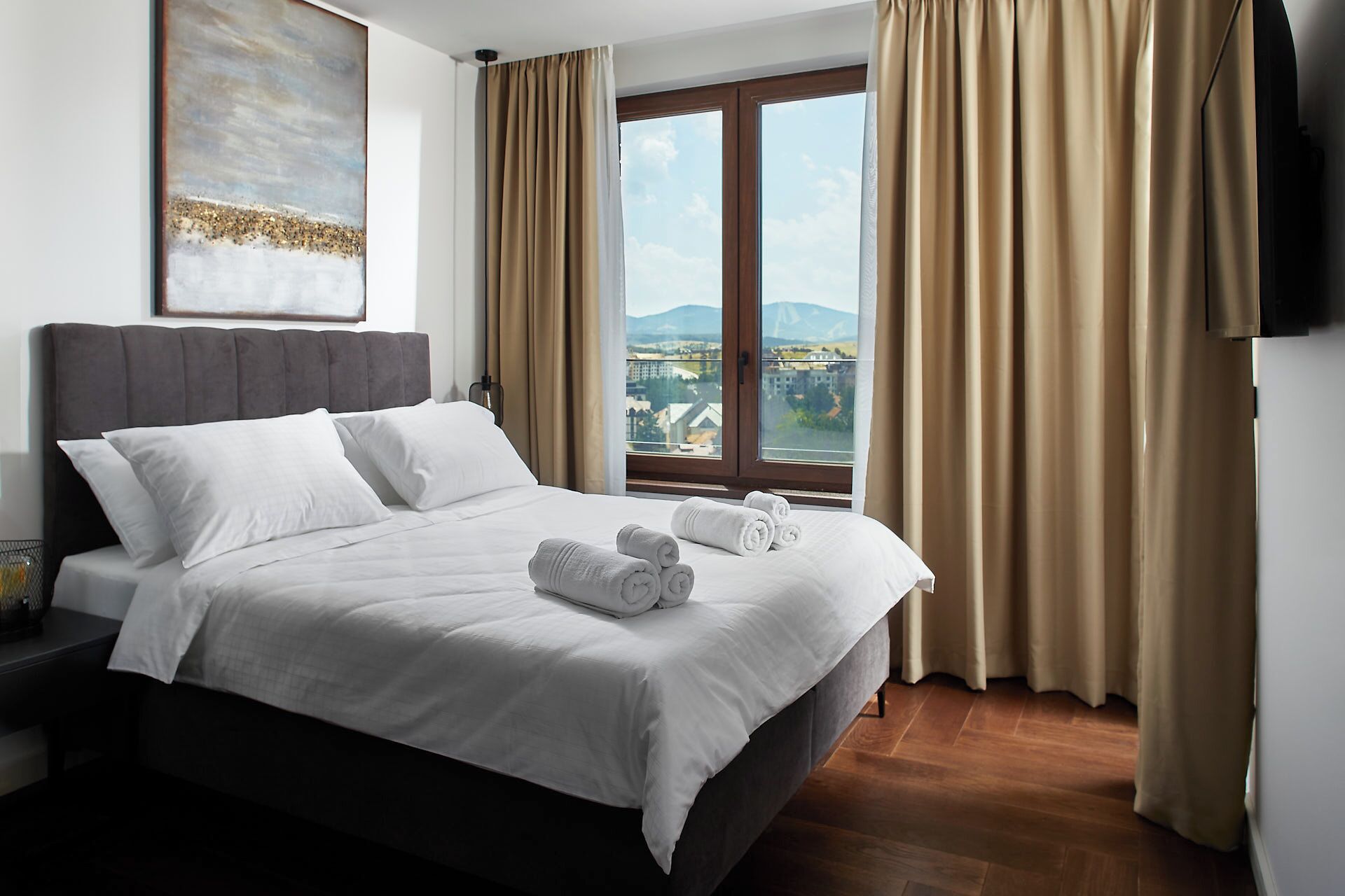 A hotel room with a bed, a view of a cityscape, and a painting on the wall.
