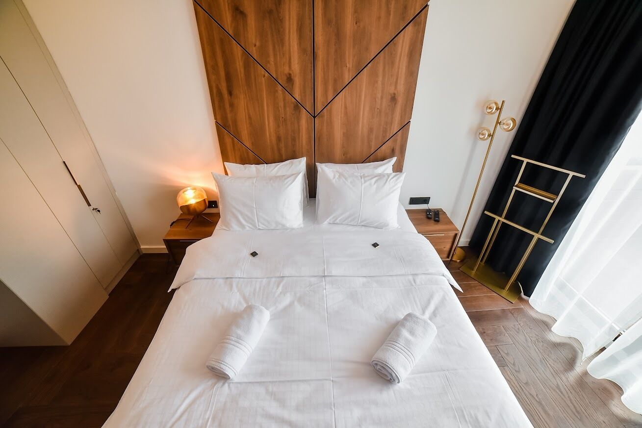 Ein ordentlich bezogenes Bett mit weißen Leinen und zwei aufgerollten Handtüchern, ein Holz-Kopfschild, eine Nachttischlampe und ein schwarzer Vorhang.