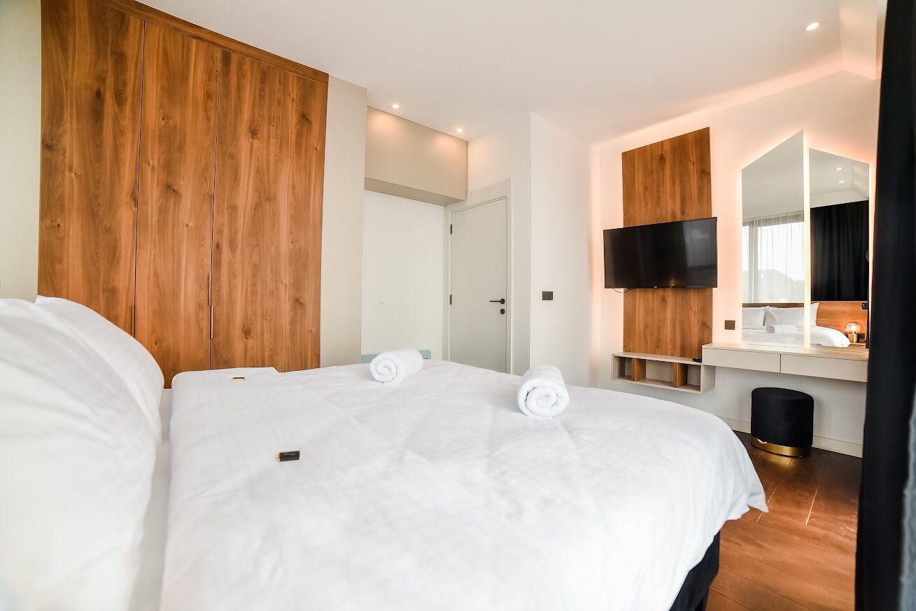 A modern hotel room with a wooden headboard, a flat-screen TV, a bedside table, and a large mirror.