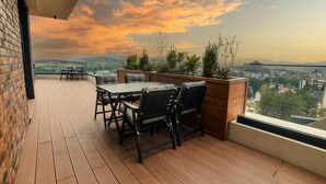 Een dak terras met een houten terras, een tafel met stoelen en uitzicht op de stadsbelangstelling bij zonsondergang.