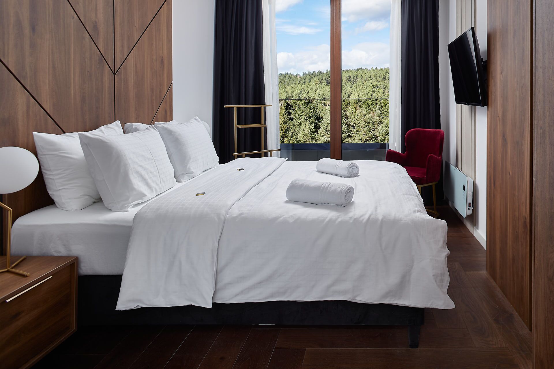 A neatly made bed with white linens, a red chair, and a TV mounted on the wall.