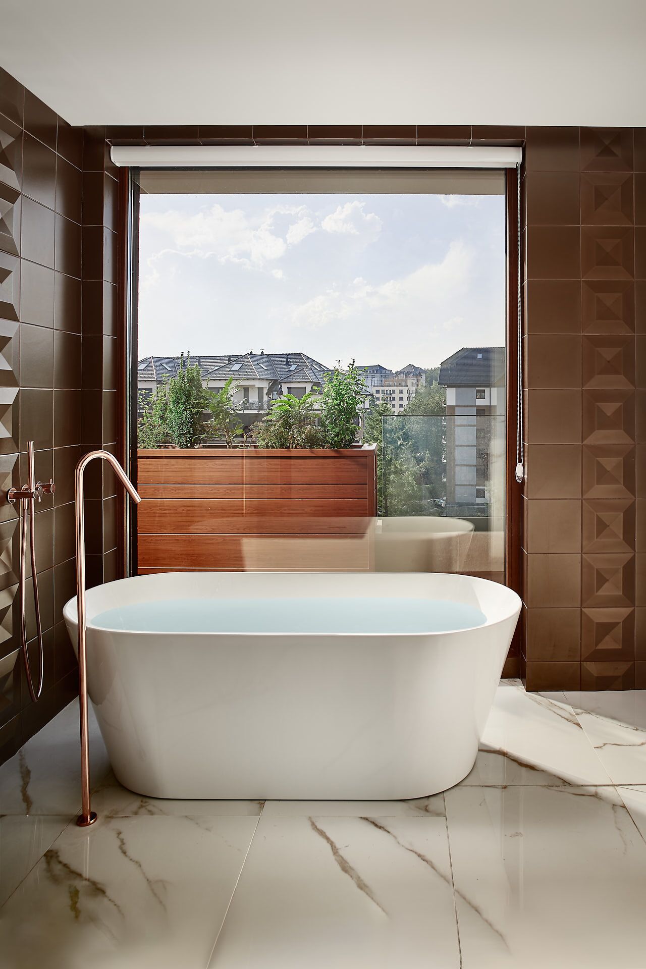 A modern bathroom with a freestanding bathtub, a wooden bench, and a view of buildings through a large glass door.