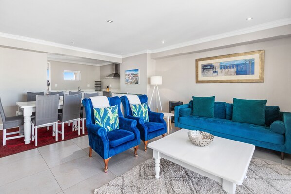 Una sala moderna con un sofá azul, dos sillones azules, una mesa de centro blanca y un comedor con un tapete rojo.