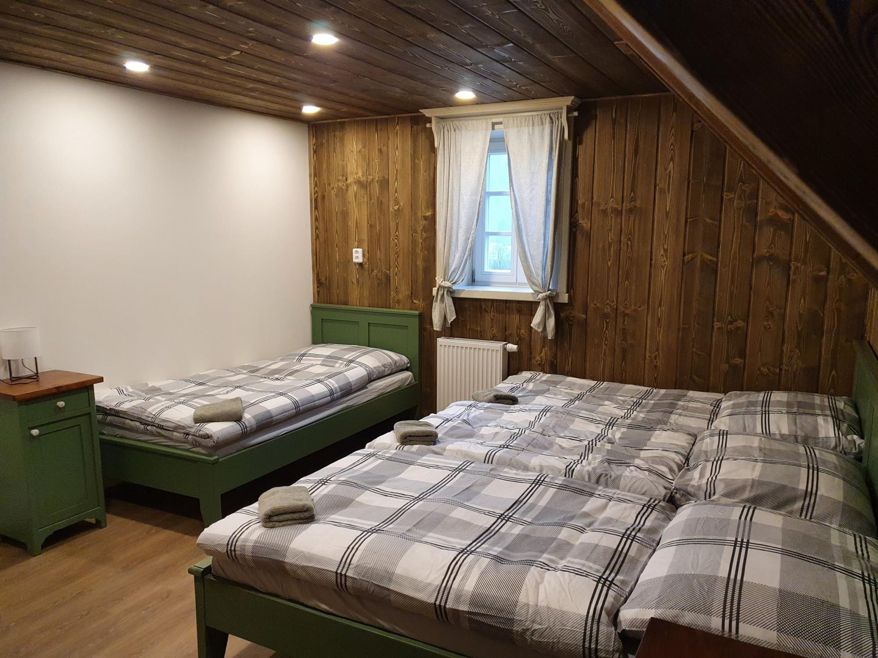 Habitación con dos camas, paredes de madera, una ventana con cortinas, un radiador y una mesita con lámpara.