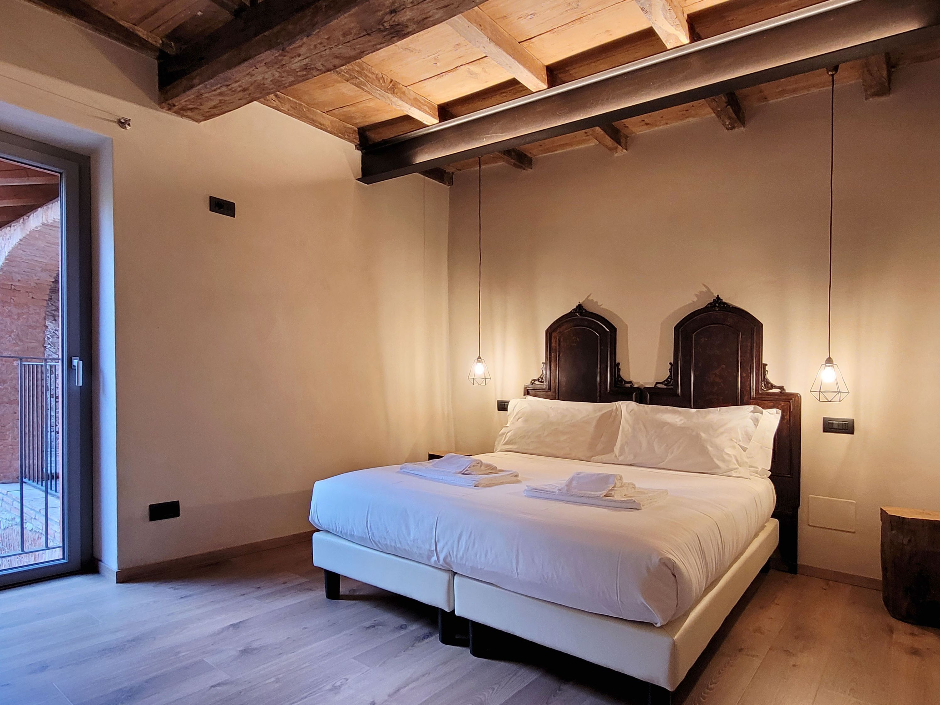 Quarto com teto de madeira, uma cama grande com roupa de cama branca, duas luzes fixadas na parede e uma mesa de cabeceira de madeira.