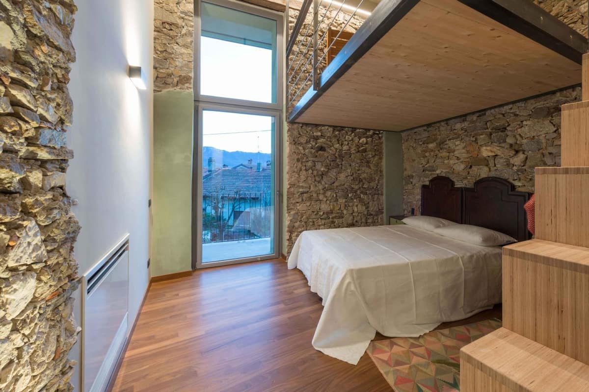 A bedroom with a stone wall, a bed, a wooden staircase, and a view through a large window.