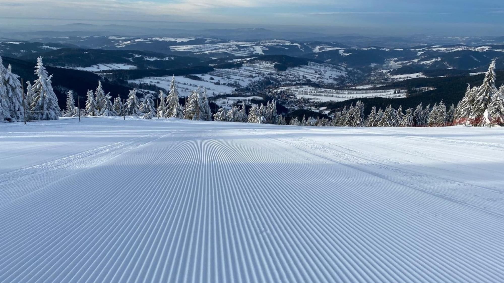 A snow-covered ski slope with a clear view of distant mountains and trees.